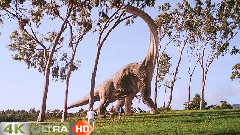 A still from the opening of the film "Jurassic Park", showing a brachiosaurus among trees and some people running towards it